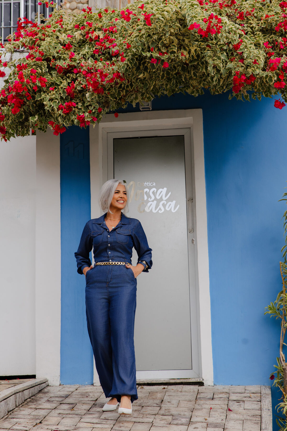 Fundadora Dri Moren na porta da Estética Nossa Casa, com flores de trepadeira acima da entrada